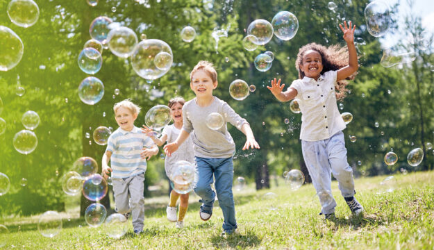 Symbolbild Kindergeburtstag. Kinder spielen draußen mit Seifenblasen.