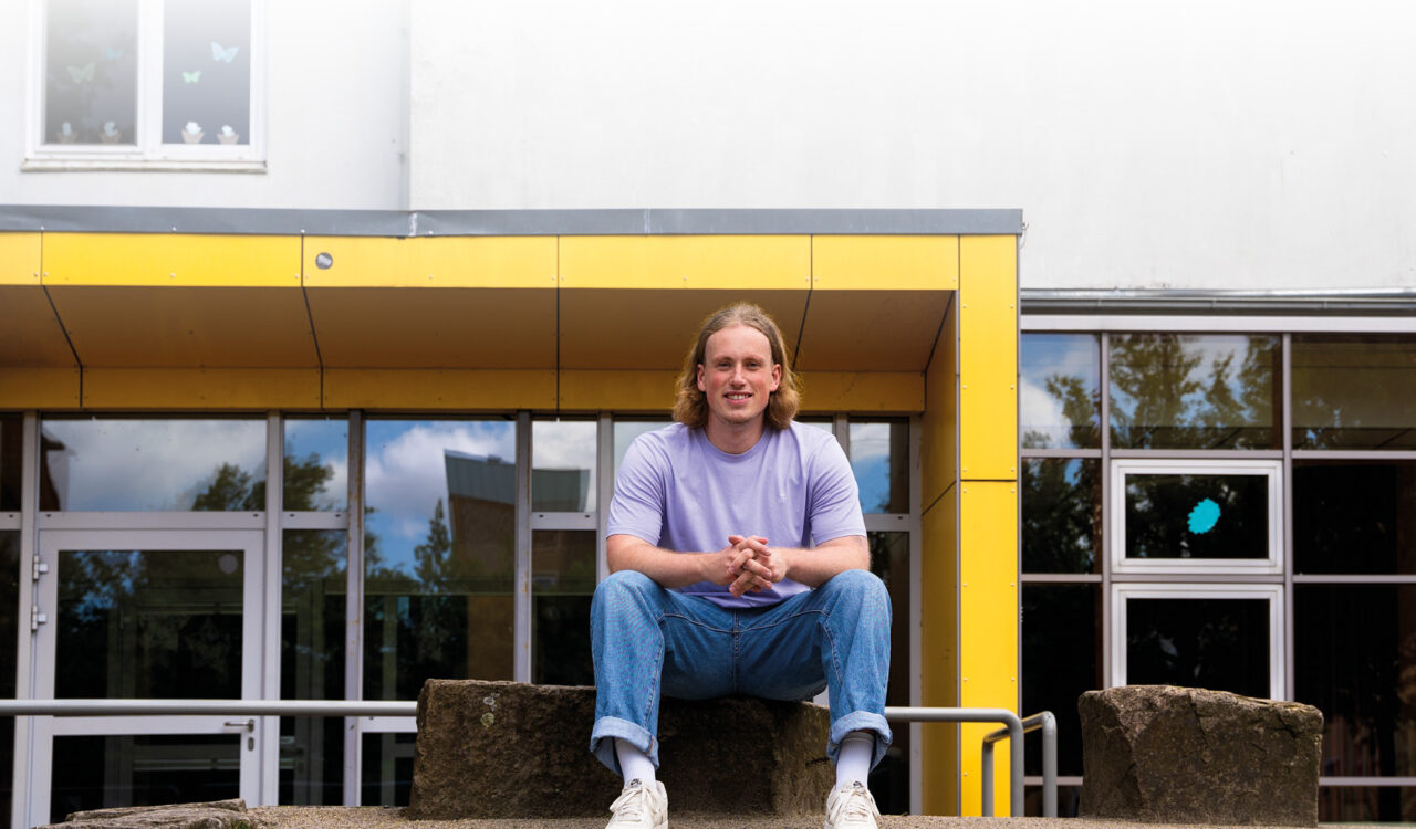 Der junge Lehrer Lukas Trumm sitzt lächelnd auf einer Stufe vor dem Eingang zur Astrid-Lindgren-Schule in Bremerhaven