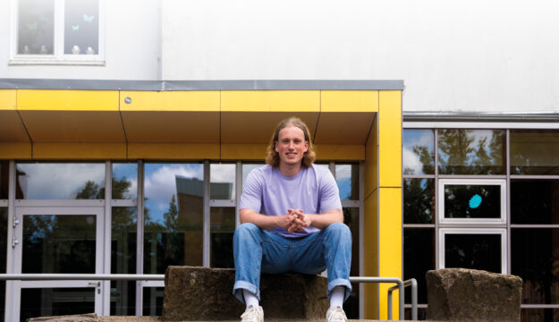 Der junge Lehrer Lukas Trumm sitzt lächelnd auf einer Stufe vor dem Eingang zur Astrid-Lindgren-Schule in Bremerhaven