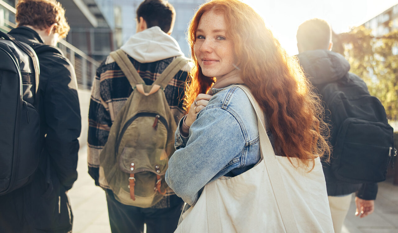 Eine junge Studentin mit Tragetasche blickt inmitten einer Studierendengruppe über die Schulter